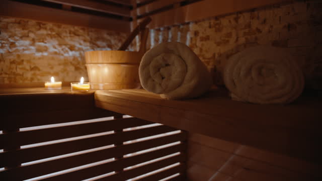 Burning Candles On A Wooden Shelf Illuminate A Serene Sauna Spa With Rolled-Up Towels Nearby