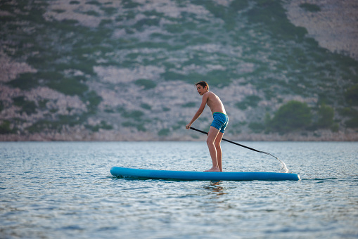 Teenage boy in blue trunks standing on inflatable board and rowing, active summer vacation at the seaside