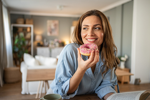 The photo shows a young woman in her cozy apartment, sitting at the kitchen island in the kitchen and indulging in her favorite book while enjoying delicious donuts