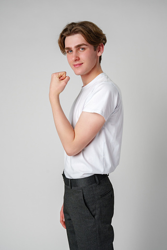 A young man with wavy brown hair presents a confident posture while displaying his flexed bicep. Dressed in a casual white t-shirt and dark pants, he exudes a relaxed yet assertive demeanor against a neutral gray backdrop, illustrating an empowering stance.