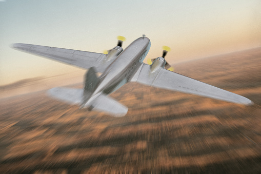 A scale model of a bare-metal DC-3 Dakota in flight, seen here flying over a desert location at dusk.