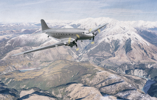 A scale model of a bare-metal DC-3 Dakota in flight, flying high above the snow-capped peaks of a mountain range.