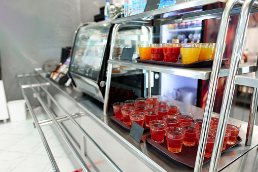 A selection of fruit juice cups, with orange and red hues, are neatly arranged on a metal serving cart, ready for patrons to enjoy in a bustling cafeteria setting. The focus on the front cups, with a blurred background, highlights the refreshment options available.