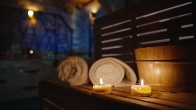 Burning Candles On A Wooden Shelf Illuminate A Serene Sauna Spa With Rolled-Up Towels Nearby