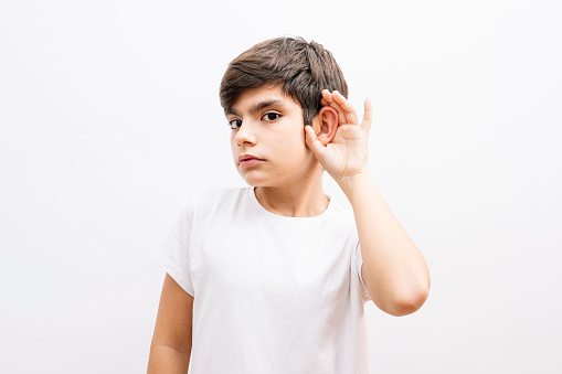 Beautiful kid boy wearing casual t-shirt standing over isolated white background with hand over ear listening an hearing to rumor or gossip. Deafness concept.