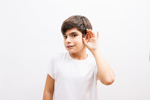Beautiful kid boy wearing casual t-shirt standing over isolated white background smiling with hand over ear listening an hearing to rumor or gossip. Deafness concept.