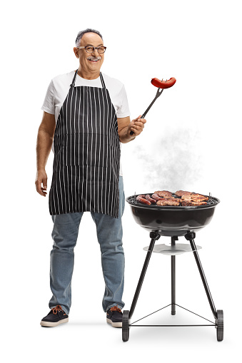 Mature man next to a portable barbecue grill holding a sausage with a fork isolated on white background