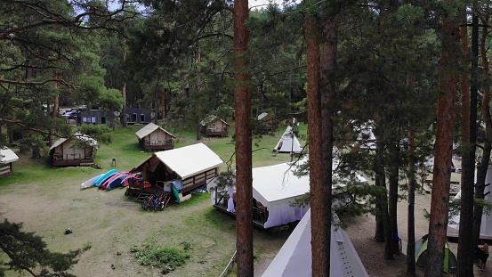 A tent camp with wigwams and bungalows, surrounded by pine trees in the forest. A SUP (stand-up paddleboard) is available for guests to enjoy the calm waters of the lake. Aerial footage. 4k