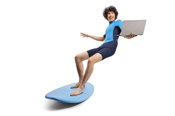 Young man in a wetsuit riding a surfboard and holding a laptop computer isolated on white background