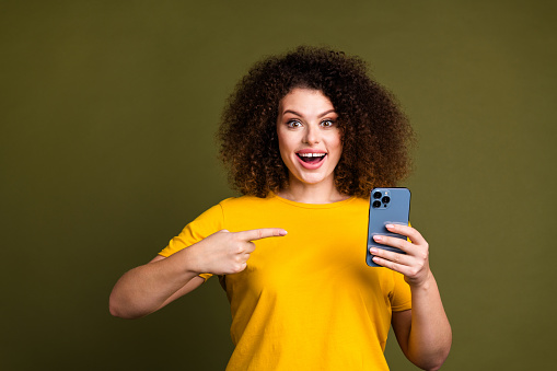 Photo of young funky amazed funny lady wearing yellow t shirt pointing finger ios smartphone update isolated on khaki color background.