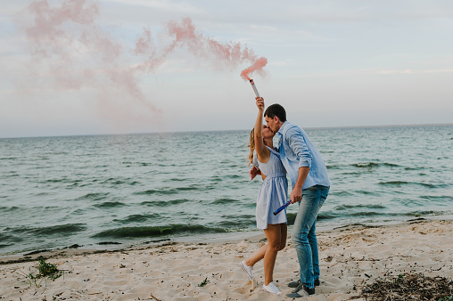 Man and woman standing and kissing on sand and holding colorful red or pink color smoke bombs on beach.  Married couple with a smoke bomb in happy summer day. Baby shower. It is a girl.