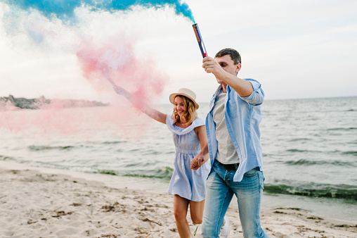 Man and woman having fun running on sand and holding colorful blue and red or pink color smoke bombs on the beach. A happy married couple holds hands smoke bomb. Baby shower. Twins:  boy and girl.