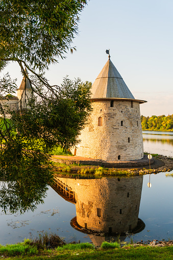 The mighty walls of an old medieval fortress, a watchtower, the rays of the dawn sun, a river near the wall, reflection in the water, reeds in the water, parks and nature, green vegetation, tree foliage, travel and tourism, autumn.