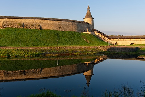 A magnificent view of the walls and watchtowers of an ancient fortress at dawn, hipped roofs, calm surface of water, reflection of buildings in the water, ancient Russian architecture, parks and nature, plants and trees, cityscape, sunlight, travel and tourism, autumn.