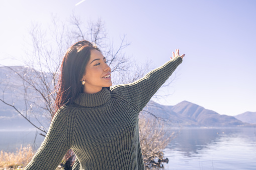 A young woman with an infectious smile, arms raised in a carefree gesture, embracing the beauty of the outdoors. Dressed in a comfortable, chunky knit sweater, she stands by a pristine lake, basking in the sun's glow. The landscape is breathtaking with the lake's shimmering surface reflecting the clear sky and the silhouette of mountains in the distance.