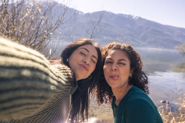 A selfie of two smiling women enjoying a sunny day by the lake. The woman in the foreground is wearing a cozy green sweater, and her friend is in a complementary green tee. Both are making happy and goofy faces, with the glistening lake and a mountain range providing a stunning natural backdrop.