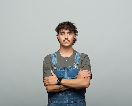 Gen z young man with curly hair and mustache wearing khaki shirt and dungarees standing with arms crossed and looking at camera. Studio shot, grey background.