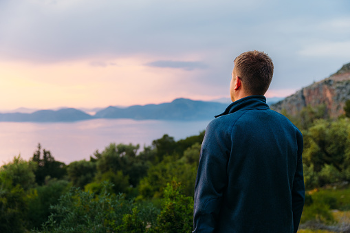 Side view of a male contemplating view from above of Mediterranean sea in pink orange sunset lights with the hills of Oludeniz in Turkey