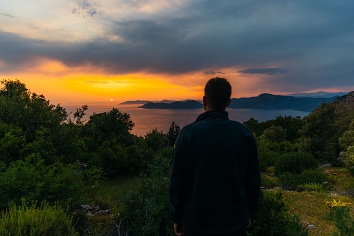 Rear view of a male contemplating view from above of Mediterranean sea in pink orange sunset lights with the hills of Oludeniz in Turkey