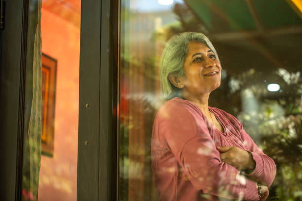 istock Thoughtful senior woman looking through window while standing at house 2148710019