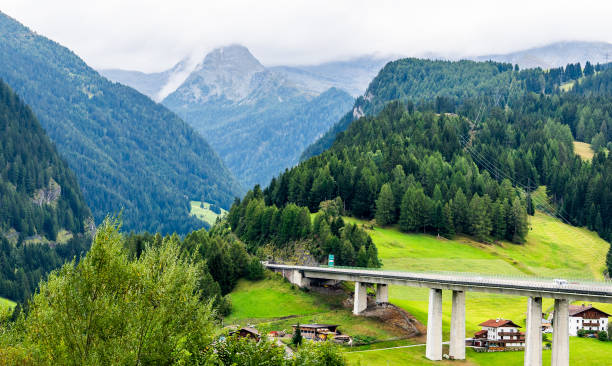 An elevated highway stretches across a verdant landscape, supported by sturdy concrete pillars. Below, a small village with traditional houses nestles amidst lush greenery. Towering mountains, their peaks shrouded in mist, provide a dramatic backdrop under an overcast sky. Shot taken in Italy.