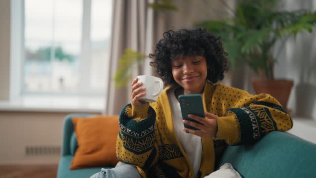 Close up young girl with phone drink tea. African American woman hold in hand use mobile phone drink coffee. Girl relax spend free spare time at modern home with phone. Communication with friends.