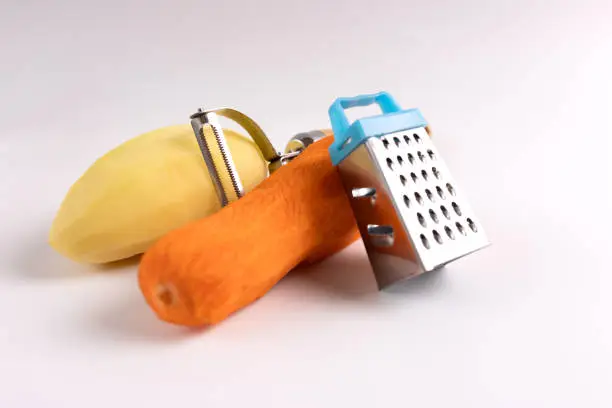 Raw carrots, potatoes, skinning knife and grater on a white background.