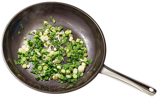 Browning chopped up young Spring Scallions in a large heavy duty non-stick ceramic coated black frying pan, isolated on white background, high angle view.