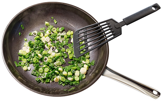Browning chopped up young Spring Scallions in a large heavy duty non-stick ceramic coated black frying pan, stirred with black plastic spatula, isolated on white background, high angle view.