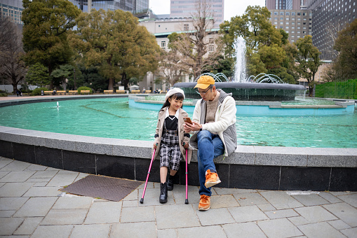 Little girl with crutches sitting with father in front of fountain in public park