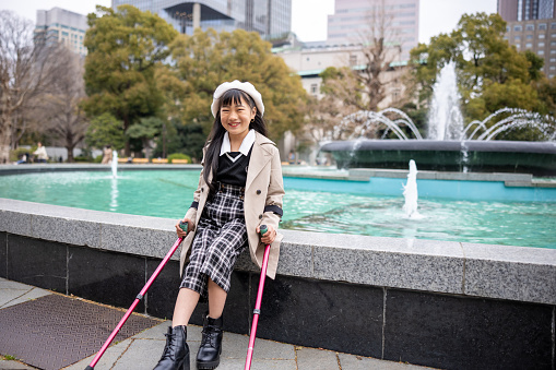 Portrait of happy little girl with crutches sitting in front of fountain in public park