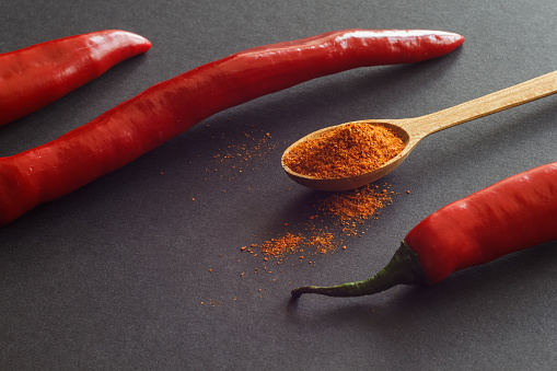A composition of pods of hot pepper and a spoon with milled pepper on a dark background