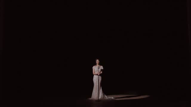 Woman in evening dress stands alone on stage near microphone. The girl speaks into the microphone. A young girl sings in a large dark hall. Elegant bride in white gown in soft light. Woman in wedding dress with glowing backdrop. Bridal beauty