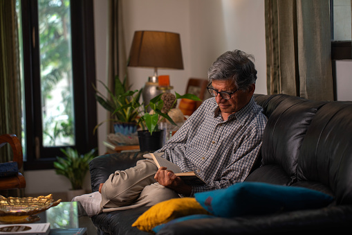 Serious old man in eyeglasses reading book with focus while relaxing on sofa in living room at home