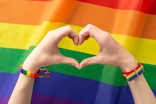 Celebrate LGBT History Month with this beautiful top-view photo of a female hand, sporting a symbolic bracelets, forming a heart with her fingers rainbow flag serving as background