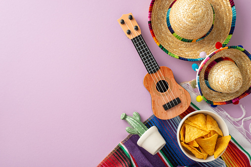 Cinco de Mayo concept. Top view picture featuring characteristic gear: sombreros, stringed vihuela, a cactus, a bright serape, set on a pastel purple field, space for text