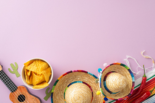A top-view snapshot for Cinco de Mayo, showing festive sombreros, a stringed vihuela, a patterned serape, hot chilli, and nachos on a pastel purple surface