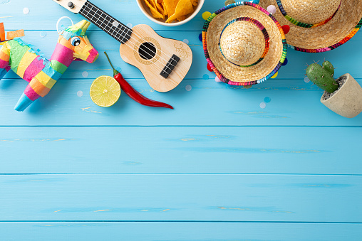 Celebrate Cinco de Mayo with this vibrant top view scene: sombreros, vihuela, cactus, lama pinata, lime, chilli pepper, nachos and confetti on blue surface. Ideal for event promos