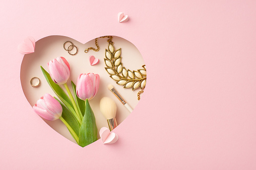 Stylish Mother's Day concept: Top view of tulips, makeup tools, gold accessories, and paper hearts in a heart-shaped frame against pastel pink background, space for greetings or adverts