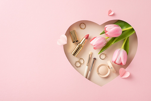 Mother's Day sophistication: Top view of tulips, makeup brushes, lipstick, eyeshadow, gold jewelry, and paper hearts in a heart-shaped frame on soft pink, with space for greetings or adverts