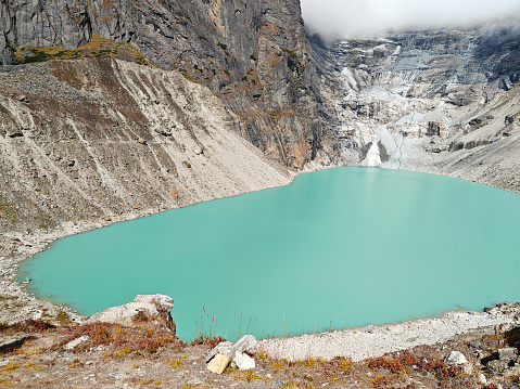 A turquoise glacial lake nestled in the Himalayan mountains of Nepal, with cloudy skies above.