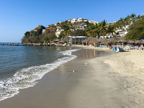 La Cruz de Huanacaxtle, Nayarit, Mexico - 2024-04-08: The serene morning atmosphere at Playa la Manzanilla in La Cruz, features gentle waves lapping the sandy shore, with a few early beachgoers enjoying the calm, and thatched-roof palapas awaiting the day’s visitors, all set against a backdrop of lush hills and luxury residences.