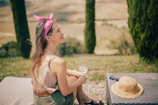 Young woman in casual attire wearing striped bandana sitting on deckchair holding glass of white wine, smiling happily enjoying scenic view whilst on vacation in countryside with blurry Tuscan landscape in background