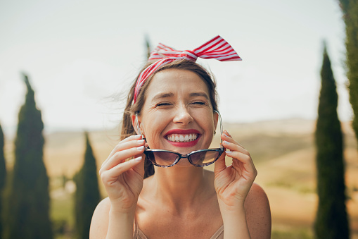 Happy young woman wearing striped bandana removing sunglasses smiling cheerfully at camera with eyes closed, enjoying cool weather and sun whilst on vacation with blurry Tuscan landscape in background
