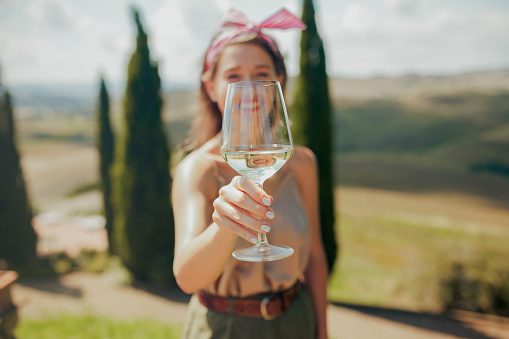 Glass of white wine held in woman's hand with painted nails, posing at camera with blurry background of woman smiling on hilltop with blurry Tuscan landscape in background