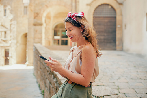 Young female tourist in casual wear sitting on stone ledge, smiling happily and sharing holiday photographs in social media on cellphone, enjoying alone time sightseeing in medieval village