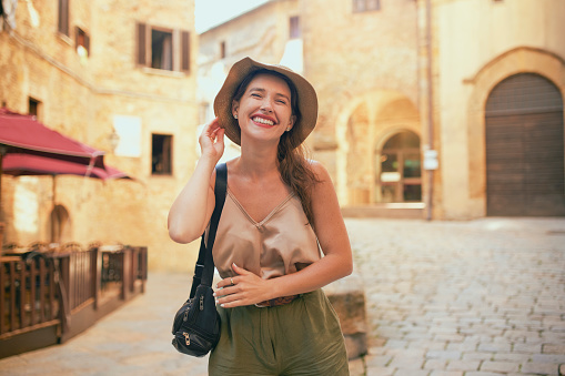 Lovely female tourist wearing casual attire and straw hat, standing on walkway surrounded with stone buildings in medieval village, smiling joyfully with closed eyes