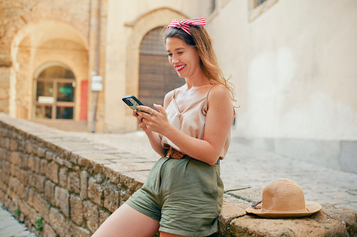 Young female tourist in casual wear sitting on stone ledge beside straw hat, smiling happily amused with video reels in social media on cellphone, enjoying alone time sightseeing in medieval village