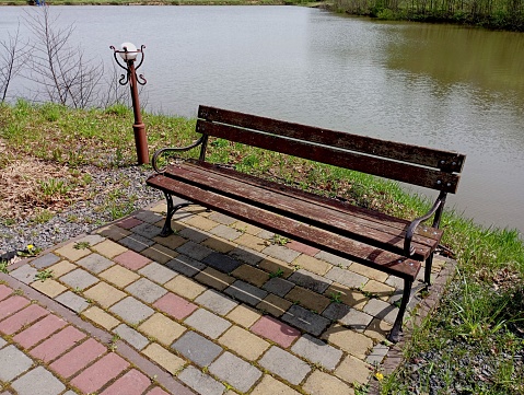 A metal bench with a wooden seat on the shore of a pond on a small cobblestone area. Next to the bench is a garden post with a lantern.