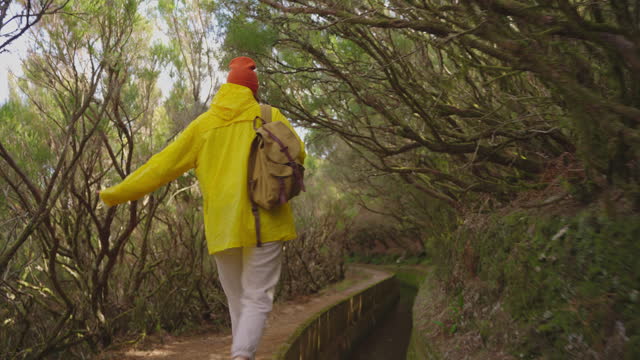 A girl in a yellow jacket travels with a backpack and visits extraordinary places and trails on the island of Madeira, Levada dos Tornos, Portugal